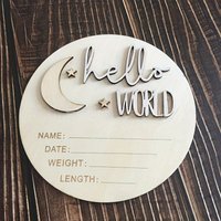 A circular wooden "Hello, World" birth announcement board with the design of a moon and stars.